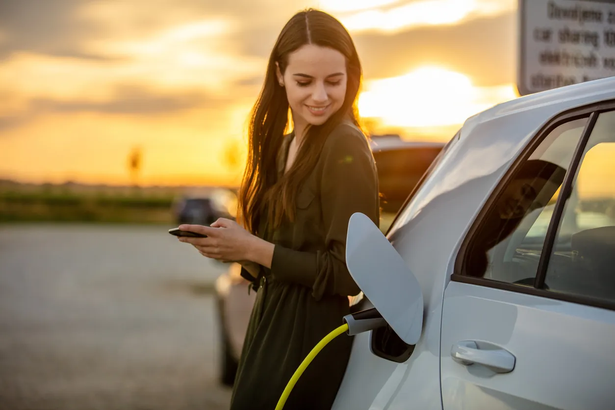 oman waiting for electric car to charge in the parking lot at sunset