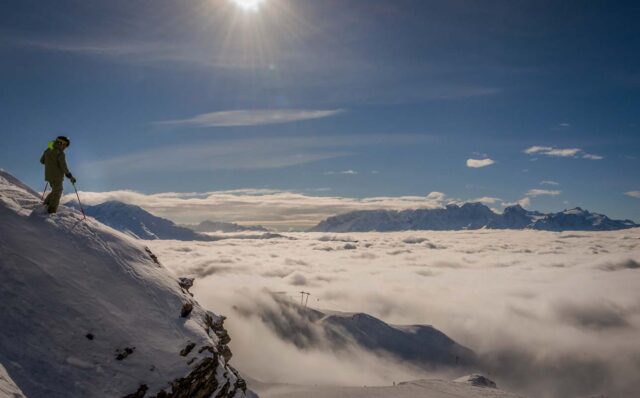 A skier in the Swiss ski resort of Verbier standing on top of a cliff above a sea of clouds, looking towards Mont Blanc and Chamonix.